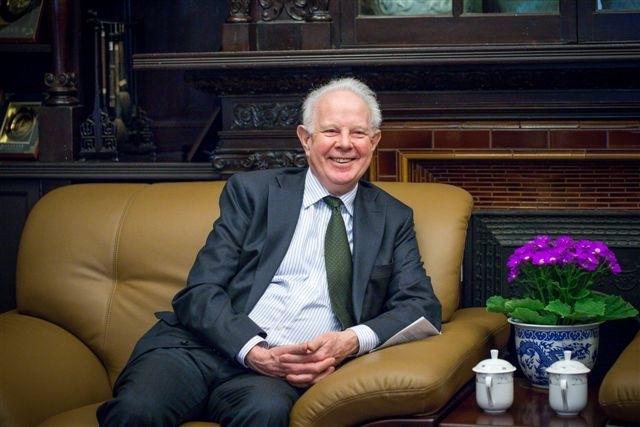 A picture of Lord Thomas sitting on a mustard sofa and smiling. There are purple flowers by the side of the sofa. Lord Thomas is dressed in a grey suit and is wearing a striped white shirt and green tie.