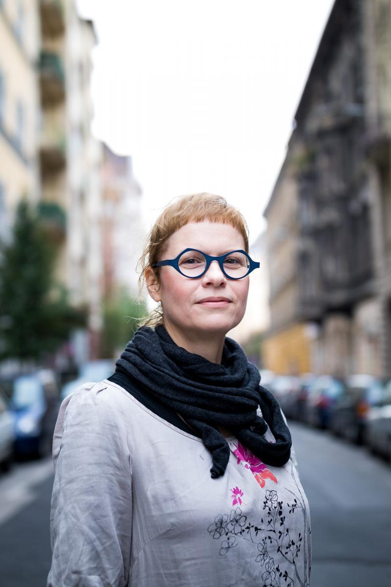 A photo of Renata Uitz, pictured in a grey shirt, black scarf and blue-framed glasses.