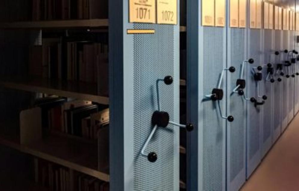 Image of a row of blue metal compact shelving units in an archive or library. The shelves have wheels for adjusting the spacing and are labeled with numbers. Some shelves are partially open, revealing books and files inside.