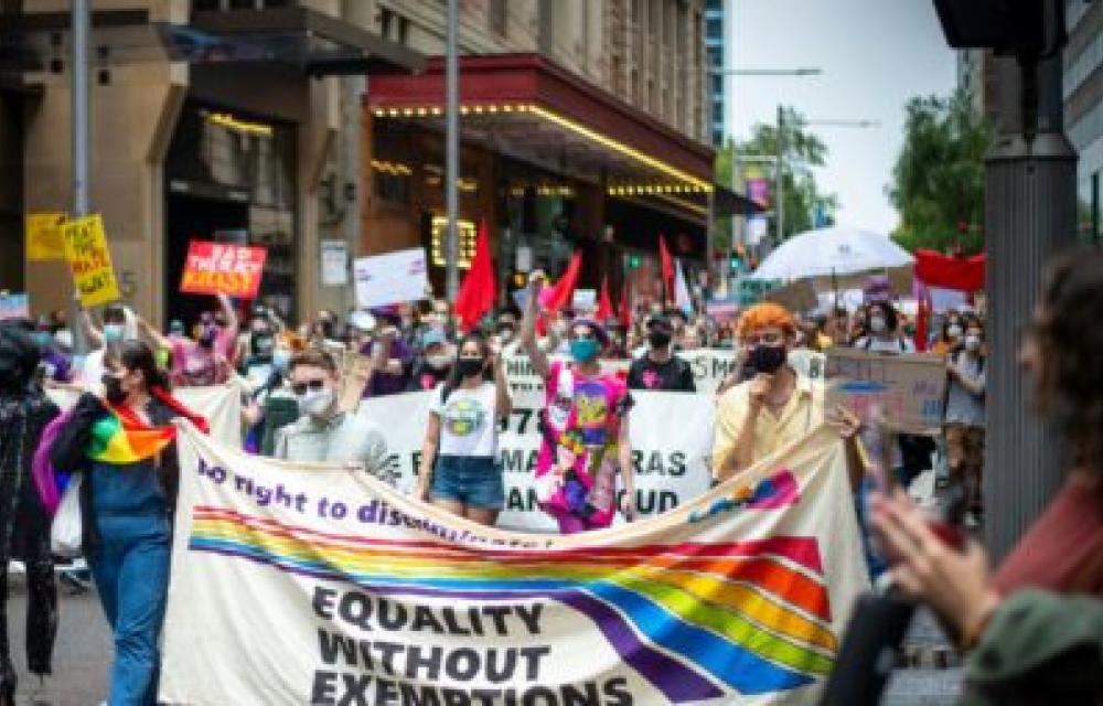 A crowd of people is marching in a city street, holding various signs and banners advocating for equality and rights. The largest banner in the foreground displays the message "Equality Without Exemptions" with a rainbow stripe, suggesting support for LGBTQ+ rights. Many participants are wearing masks, indicating a contemporary setting during a time when health precautions are necessary.