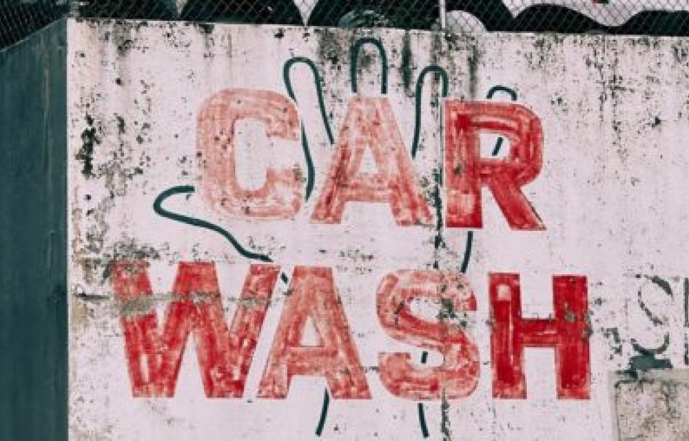 Weathered sign reading 'CAR WASH' in large red letters on a worn concrete wall with a faint outline of a hand in the background, showing signs of dirt and grime from prolonged exposure to the elements.