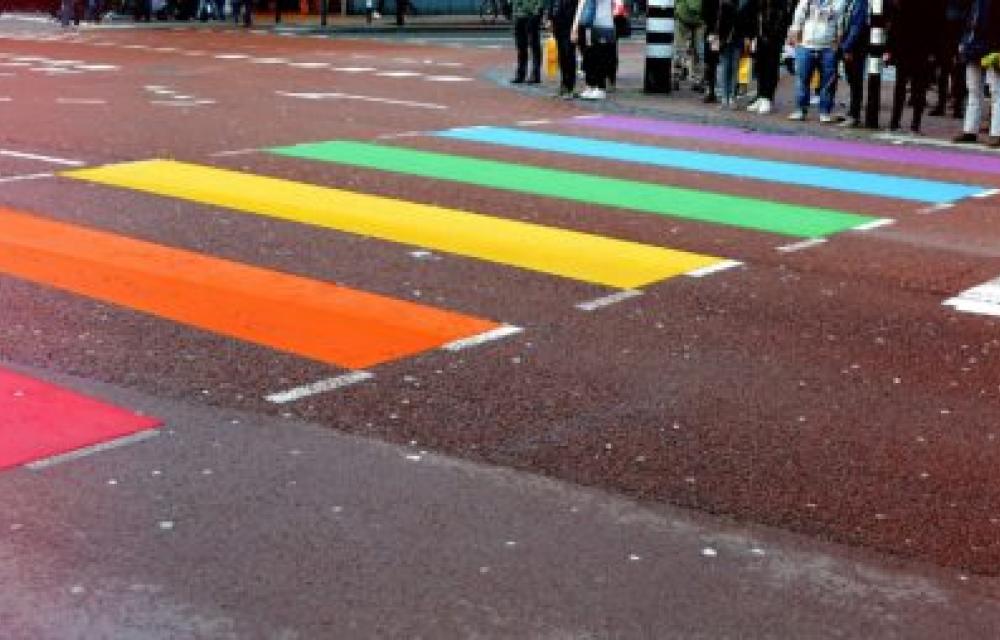 A crosswalk painted with rainbow-colored stripes, representing the LGBTQ+ pride flag. Several people are waiting to cross the street in the background.